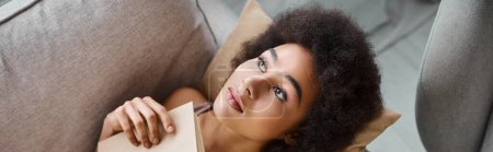 vista superior de la ropa interior de mujer afroamericana joven acostada con libro abierto en un cómodo sofá, pancarta