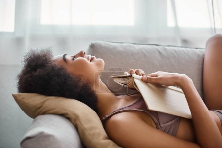Téléchargez les photos : Vue latérale de plaisir jeune femme afro-américaine sous-vêtements couché avec livre ouvert sur un canapé confortable - en image libre de droit