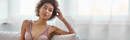 Mujer afroamericana joven reflexiva descansando en lencería en un sofá en casa, pancarta horizontal