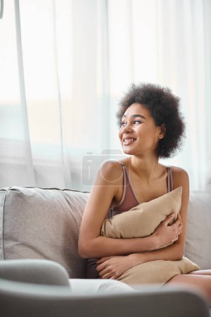 junge afrikanisch-amerikanische Frau mit lockigem Haar entspannt auf der Couch in Dessous, lächelt und schaut weg