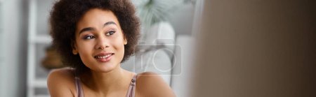 heureuse femme afro-américaine avec les cheveux bouclés relaxant sur le canapé en lingerie, bannière souriante