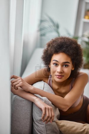 jolie femme américaine africaine avec les cheveux bouclés relaxant sur le canapé en lingerie, regardant la caméra