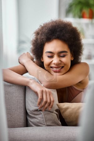 happy african american woman with curly hair relaxing on couch in lingerie, smiling with closed eyes