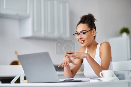 heureuse femme afro-américaine dans des lunettes de travail de la maison à distance sur son ordinateur portable, ayant appel vidéo