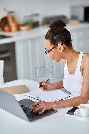 jeune femme afro-américaine dans des lunettes travaillant à distance de la maison sur son ordinateur portable, en prenant des notes