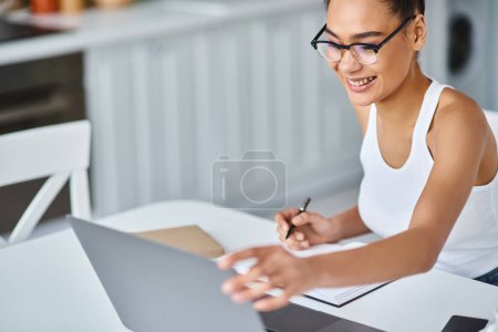 Foto de Mujer afroamericana feliz en gafas que trabajan desde casa remotamente en su computadora portátil, tomando notas - Imagen libre de derechos