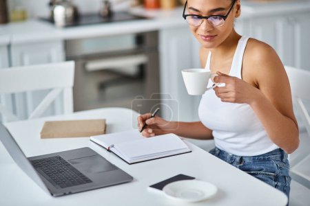 lockige Afroamerikanerin mit Brille, die von zu Hause aus ferngesteuert an ihrem Laptop arbeitet und Kaffee schlürft