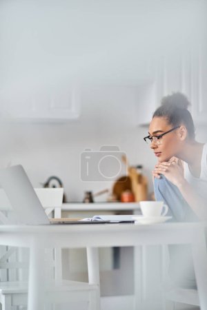 focalisée et jeune femme afro-américaine dans des lunettes de travail à distance de la maison sur son ordinateur portable