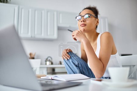 nachdenkliche afrikanisch-amerikanische Frau mit Brille, die von zu Hause aus ferngesteuert an ihrem Laptop arbeitet, Tasse Kaffee
