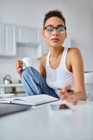 Téléchargez les photos : Femme afro-américaine bouclée dans des lunettes travaillant à la maison et atteignant smartphone flou sur le bureau - en image libre de droit