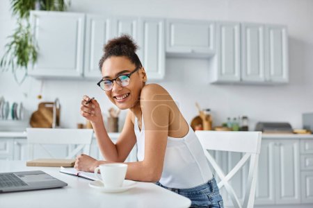 cheerful african american woman in glasses taking notes near laptop and cup of coffee, remote work