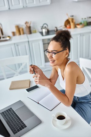 cheerful african american woman in glasses holding pen near laptop and cup of coffee, remote work