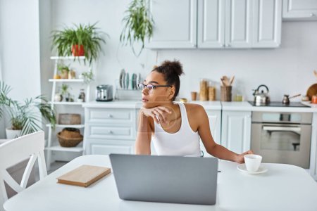 Foto de Mujer afroamericana pensativa en vasos sentados en el escritorio con computadora portátil y taza de café, trabajo remoto - Imagen libre de derechos