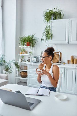 Photo for Cheerful african american woman in glasses holding cup of coffee near laptop on desk, remote work - Royalty Free Image
