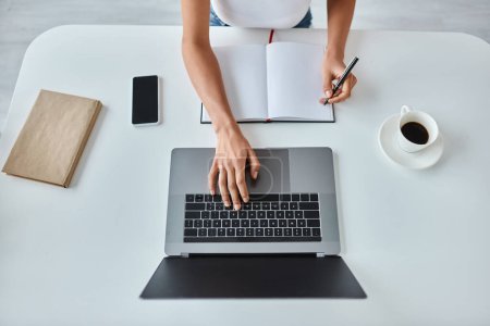 Téléchargez les photos : Vue du haut de la femme afro-américaine avec stylo à la main à l'aide d'un ordinateur portable à côté de la tasse de café sur le bureau - en image libre de droit