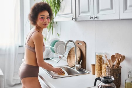 Téléchargez les photos : Femme afro-américaine gaie en lingerie avec des patchs pour les yeux plaque de lavage avec éponge dans la cuisine - en image libre de droit