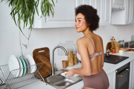 dreamy african american woman in lingerie with eye patches washing plate with sponge in kitchen