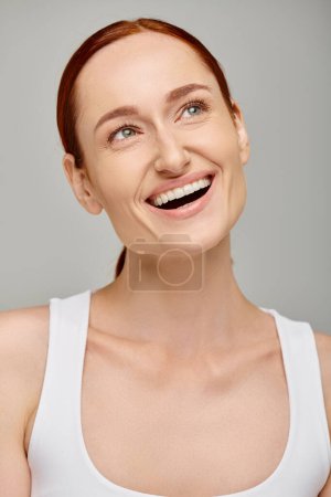 Téléchargez les photos : Heureuse rousse femme en débardeur exsudant sourire joyeux et sain sur fond gris, détournant les yeux - en image libre de droit