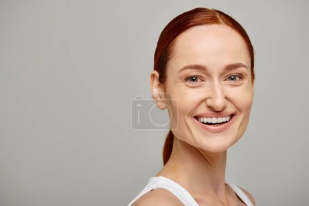 Téléchargez les photos : Femme contente et rousse en débardeur blanc avec un sourire tendre et sain sur fond gris - en image libre de droit