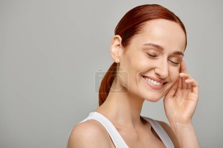 Téléchargez les photos : Femme heureuse et rousse en débardeur blanc avec un sourire tendre et sain sur fond gris - en image libre de droit