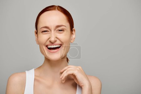 Téléchargez les photos : Femme heureuse et rousse en débardeur blanc avec un sourire sain sur un fond gris, rire - en image libre de droit