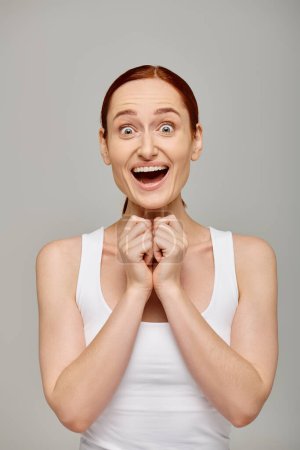 Téléchargez les photos : Femme étonnante et rousse en débardeur blanc avec un sourire sain sur un fond gris, rire - en image libre de droit