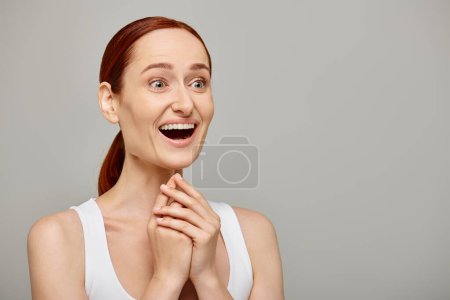 Téléchargez les photos : Femme étonnée et rousse en débardeur blanc avec un sourire sain sur fond gris, rire - en image libre de droit