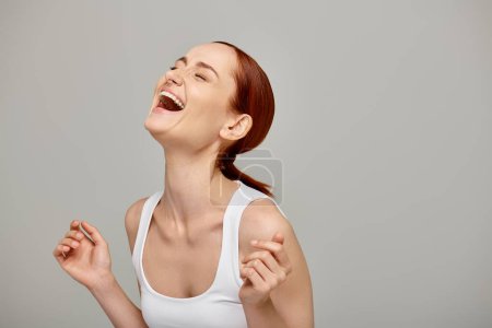 Téléchargez les photos : Femme gaie et rousse en débardeur blanc riant un fond gris, sourire radieux et sain - en image libre de droit