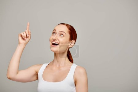 cheerful and redhead woman in white tank top pointing with finger and looking up on grey backdrop