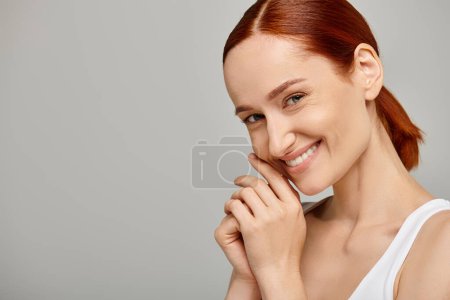 mujer feliz y pelirroja en camiseta blanca sonriendo y mirando a la cámara sobre fondo gris