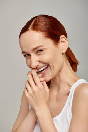 happy and redhead woman in white tank top laughing and looking at camera on grey background