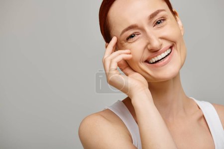 Téléchargez les photos : Femme gaie et rousse en débardeur blanc regardant la caméra sur fond gris, sourire sain - en image libre de droit