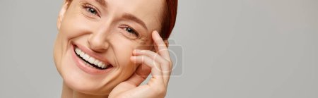 banner of cheerful and redhead woman looking at camera on grey background, healthy smile