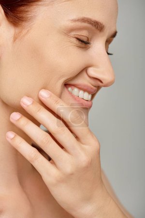 close up of joyful redhead woman with closed eyes and healthy smile touching skin on grey backdrop