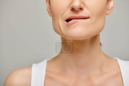 cropped view of pensive woman in white tank top biting lip, embodying concern on grey background