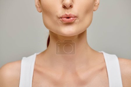 Ausgeschnittene Ansicht einer Frau in ihren 30er Jahren, die auf einem neutralen grauen Hintergrund die Lippen zusammenpustet und