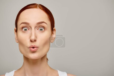 surprised young woman in white tank top looking at camera with wide eyes on grey background, wow