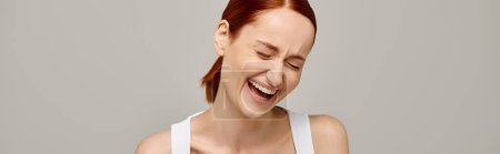 mujer feliz y pelirroja en camiseta blanca riéndose con los ojos cerrados sobre fondo gris, pancarta