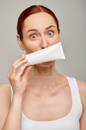 Redhead young woman in 30s covering her face with body lotion in tube on grey background, skincare