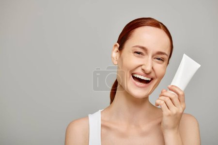 Redhead young and cheerful woman in 30s holding tube with body lotion on grey background, skincare