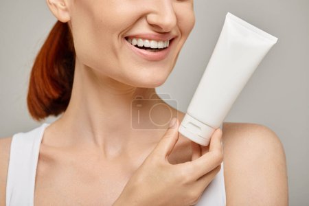 cropped view of cheerful woman with red hair holding tube with body lotion on grey background