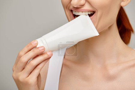 cropped view of cheerful woman with red hair biting tube with body lotion on grey background