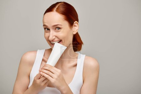 mujer alegre con el pelo rojo mordiendo tubo con loción corporal sobre fondo gris, concepto de cuidado de la piel