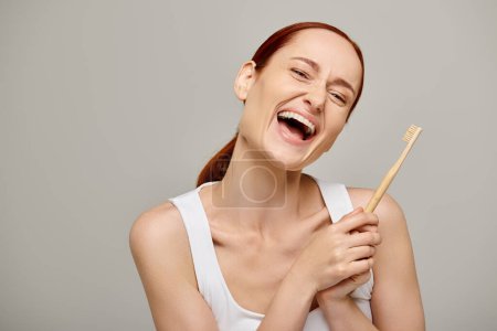 happy redhead woman in tank top holding wooden toothbrush and smiling at camera on grey background