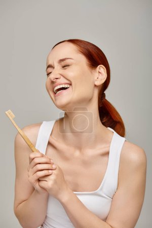 happy redhead woman in tank top holding wooden toothbrush and smiling at camera on grey backdrop