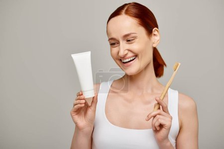 cheerful redhead woman in tank top holding toothpaste and toothbrush and smiling at camera on grey