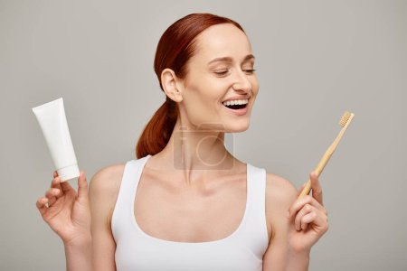 excited redhead woman in tank top holding toothpaste and toothbrush and smiling at camera on grey