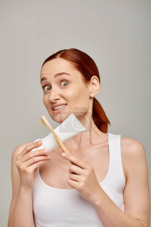 confused redhead woman in tank top holding toothpaste and toothbrush and looking at camera on grey