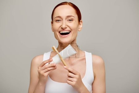 amazed redhead woman in tank top holding toothpaste and toothbrush and smiling at camera on grey