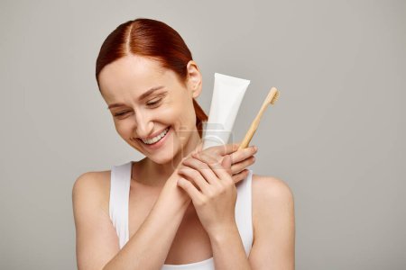 Téléchargez les photos : Joyeuse femme aux cheveux roux tenant dentifrice et brosse à dents et souriant à la caméra sur gris - en image libre de droit